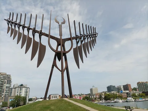 The tall rust-brown Spirit Catcher sculpture with its feather-like wings stands on a grassy hill at Barrie's lakefront. Boats sit in the marina on Kempenfelt Bay, with downtown Barrie's towers behind.