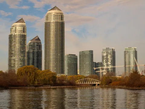 Tall round glass condo towers with pointed tops at Humber Bay Shores in Etobicoke rise behind golden autumn trees and a small white arched footbridge, with their reflections in the calm water of Humber Bay Park.