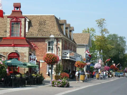 Sunny summer afternoon on Main Street Unionville in Markham. On the left are the red-trimmed brick Old Firehall with its bell tower and a mansard-roofed Victorian storefront. Hanging baskets of red and pink flowers, Canadian flags and green patio umbrellas line the brick sidewalk, which runs toward tall trees.