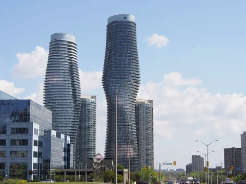 The two curving Absolute World "Marilyn Monroe" condo towers rise over Mississauga City Centre against a blue sky with white clouds, with glass office buildings on the left and a tree-lined road on the right.