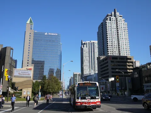 Looking north up Yonge Street in North York City Centre on a sunny day: a blue glass office tower with a green-pointed spire stands on the left, tall white condo towers rise on the right, and a red-and-white TTC bus drives toward the camera.