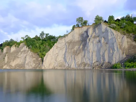 The Scarborough Bluffs in summer: towering pale clay cliffs crowned with green trees, reflected in the calm water of Lake Ontario at Bluffer's Park under a soft blue sky.