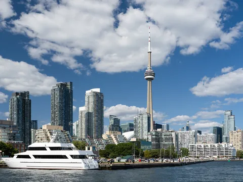 The CN Tower rising above downtown Toronto's glass condo towers under a blue summer sky with white clouds, with a white yacht docked on the harbour waterfront in front